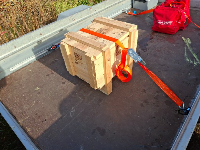 Trälåda säkrad med orange spännband på ett släpflak, bredvid röd väska. Flaket visar spår av kritmärken och vegetation i bakgrunden.