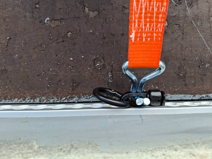 Orange spännband säkrat i metallfäste monterat på brun yta.