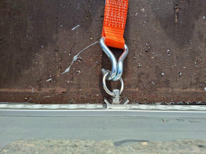 Orange spännband fäst i en metallögla på brun yta, med en monterad skena synlig nedanför.