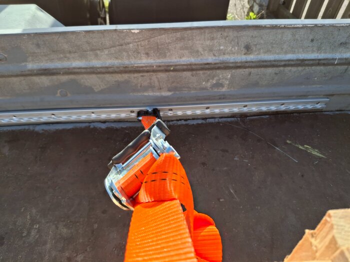 Orange spännband fastsatt i metallfäste på flak med monterad skena i bakgrunden.