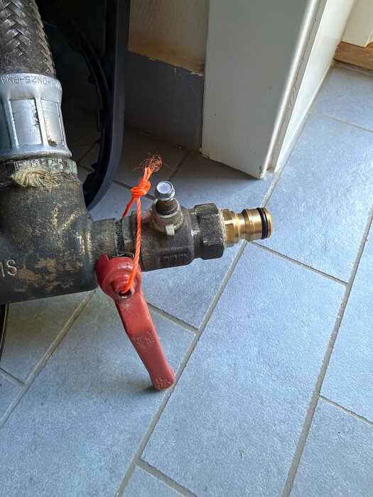 Ventil med rött handtag och orange snöre på ett grått klinkergolv, ansluten till en metallslang.