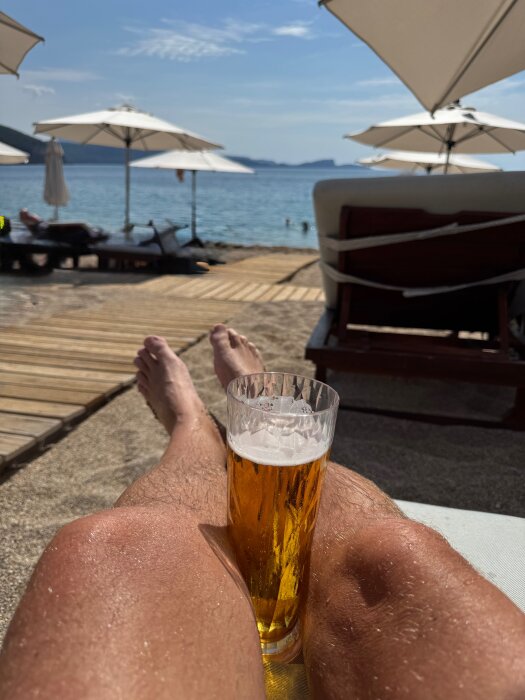 Strandvy med ett glas öl vilande på knän, parasoller i bakgrunden och havet i horisonten.