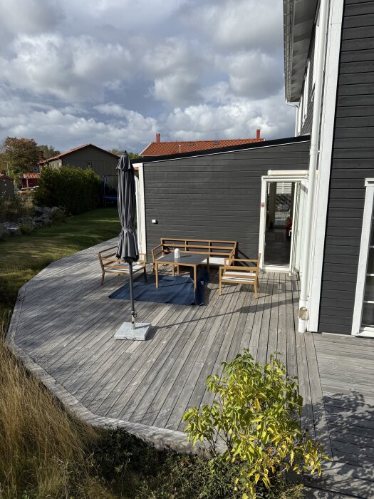 Utomhusaltan vid ett gråmålat hus, trämöbler och ett hopfällt parasoll syns på altanen, lummiga buskar runtomkring.