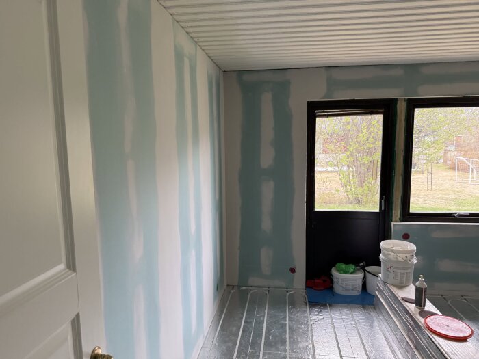 Under renovering av sovrum, delvis målad vägg, spackelmaterial och verktyg synliga, fönster med utsikt över trädgård i bakgrunden.