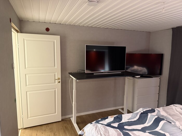 Sovrum med vit dörr, två TV-apparater på ett skrivbord, och trämönstrat golv. En säng med täcke syns delvis i förgrunden.