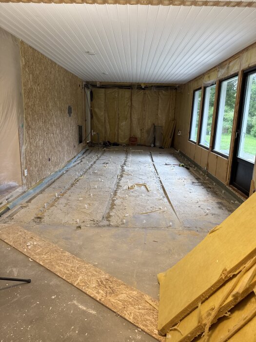 Renovering pågår i ett rum, med rivna väggar och exponerat golv. Isolering och byggmaterial syns, samt plast som täcker väggarna.