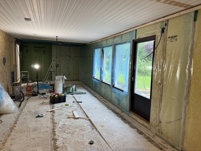 Renovering av rum med plastföreslade väggar och verktyg på golvet. Fönster täckta med blå plast; en bygglampa lyser upp rummet.