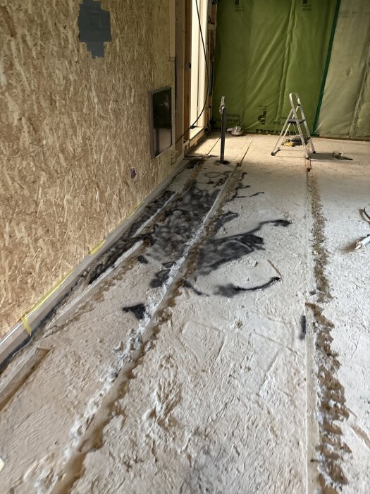 Rivningsarbete pågår med borttaget golv, blottade rör och plastade väggar i ett hus. Stege och verktyg synliga i rummet.
