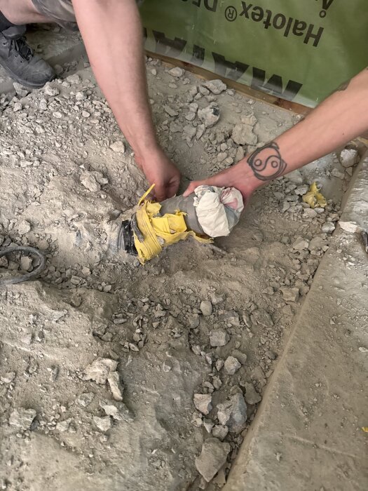 Man arbetar med ett rör i ett rivningsprojekt, omgivande golv av grus och betong. En arm med tatuering syns.
