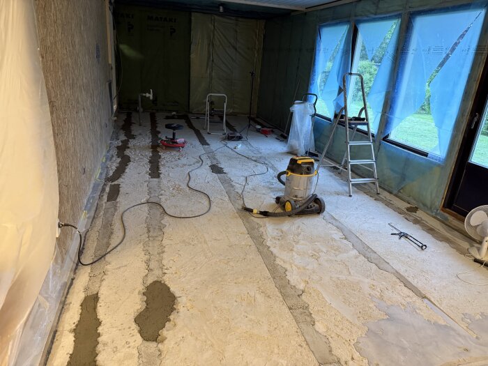 Renoveringsrum med borttagen vägg och golv, verktyg, stege och dammsugare, golv täckt med plast och isolering på väggar och fönster.