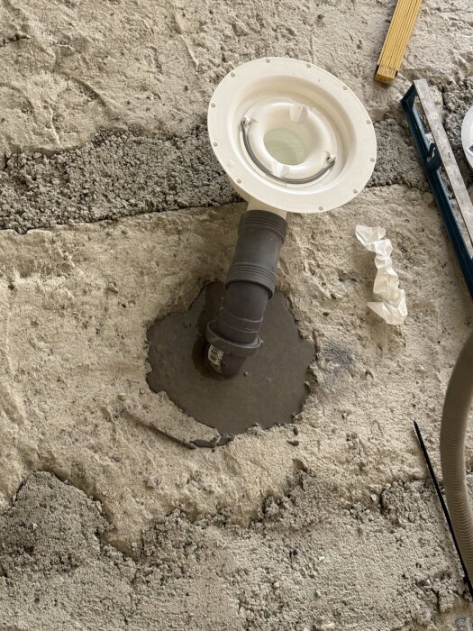 Avloppsrör och golvbrunn under renovering, omgärdat av ojämnt betonggolv. En del av rör till duschavlopp har förberetts.
