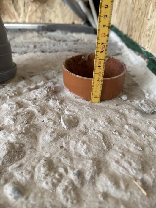 Mätrulle placerad i rödbrun muff för avloppsrör på ojämnt betonggolv, visar djup under renovering för att flytta röret.