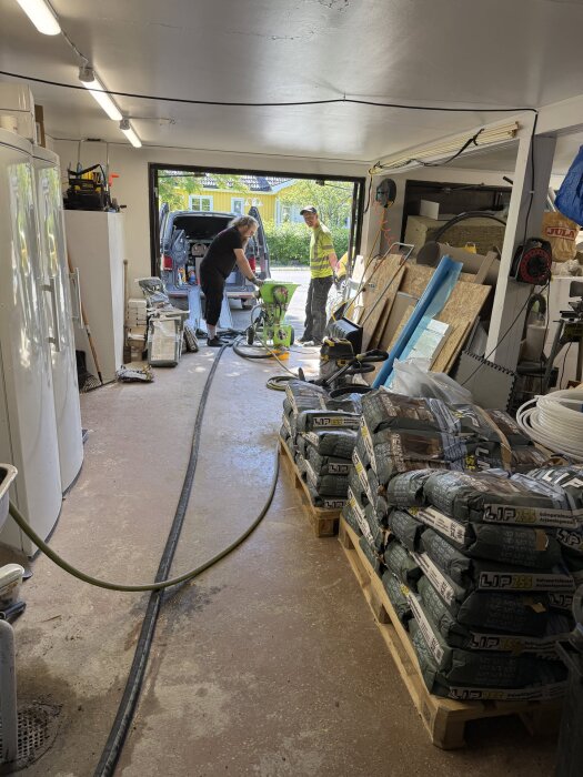 Två personer arbetar i en garage med pumputrustning, omgivna av säckar med byggmaterial på pallar och verktyg. En öppen dörr leder ut till en trädgård.