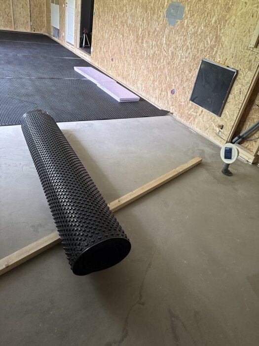 Svart rulle av Jape ventgolv ligger på ett betonggolv, omgiven av OSB-väggar och byggmaterial.