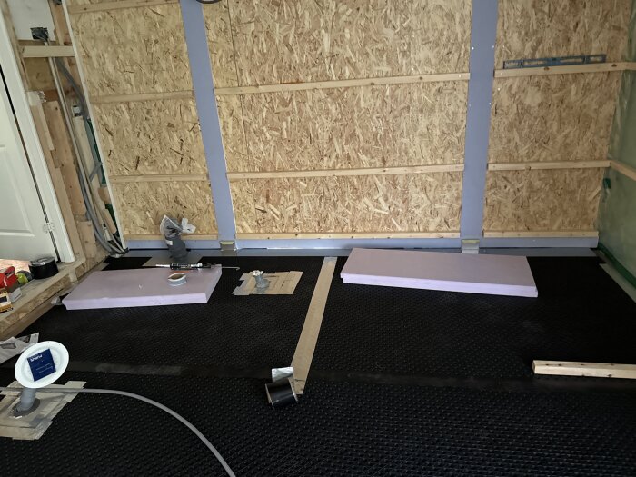 Byggarbetsplats med förberedelse för isolering, träreglar på väggen och golv med underlag av svart plast och verktyg i förgrunden.