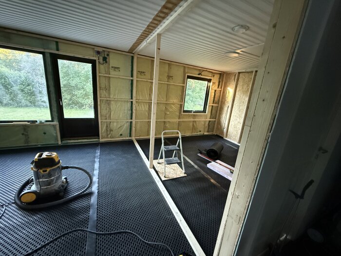 Under renovering av rum med träreglar, ventilerad golvmatta och verktyg; stege och dammsugare synliga. Skog utanför stora fönster.
