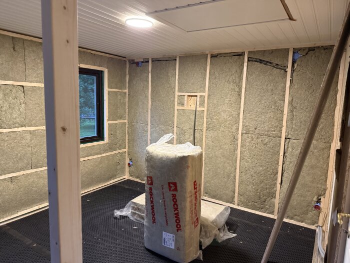 Rum under renovering med isolering på väggarna och en rulle med Rockwool-isolering på golvet, oisolerat fönster och synligt ventgolv.