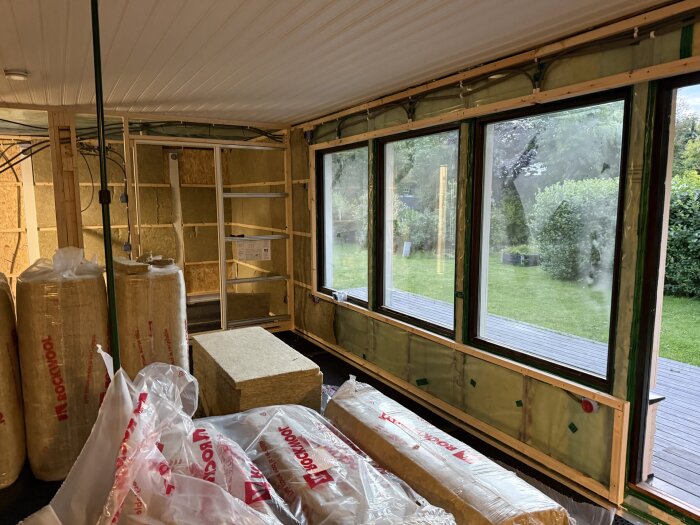 Rum under renovering med isolering och plast insatta i väggarna, stora fönster mot en grön trädgård utanför.