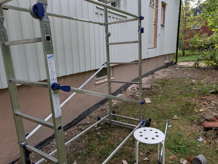 Byggnadsställning och pall utanför ett hus under renovering, med träpaneler och gräs på marken.