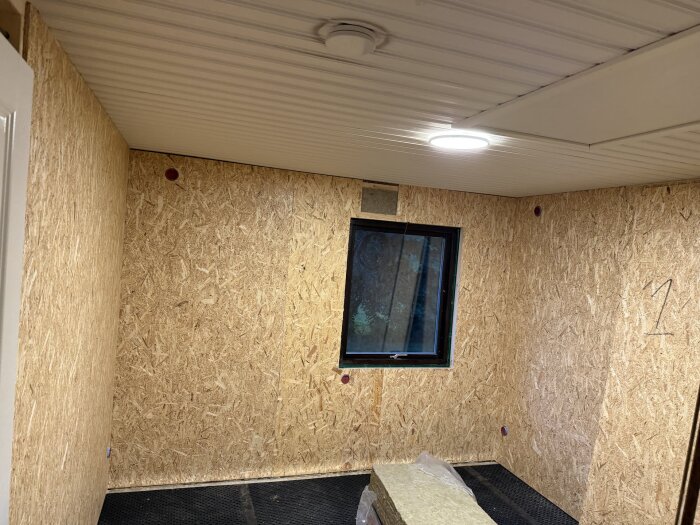 Nyrenoverat rum med OSB-skivor på väggarna, isoleringsmaterial på golvet, ett fönster mot utsidan och taklampa i tak av vit panel.