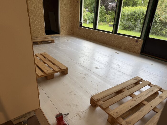 Rum under renovering med vita isoleringsplattor på golvet, OSB-skivor på väggarna och träpallar. Stora fönster med utsikt mot trädgård.