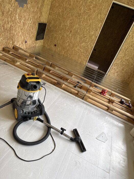 Verktyg och dammsugare i rum med plywoodväggar under renovering, med staplade byggmaterial och öppning till annat rum.