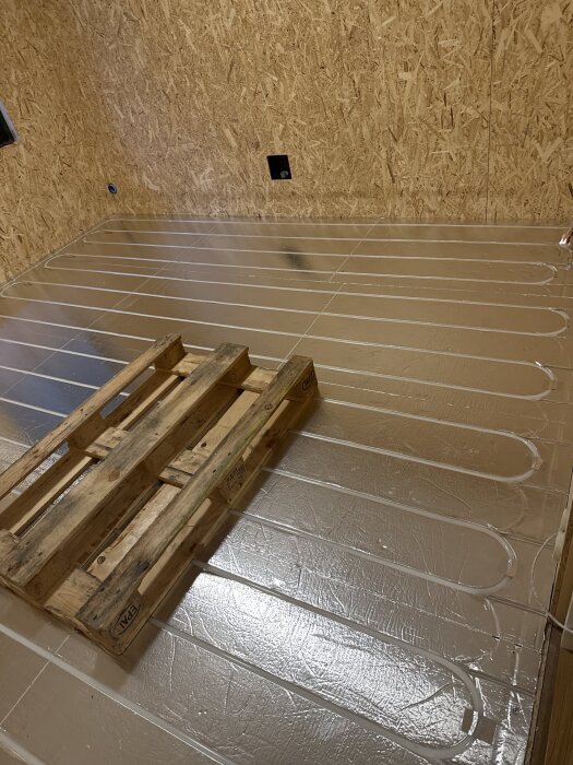 Golvisolering med 70mm cellplast, spår för golvvärmeslangar synliga. OSB väggar i bakgrunden och en träpall på isoleringen.