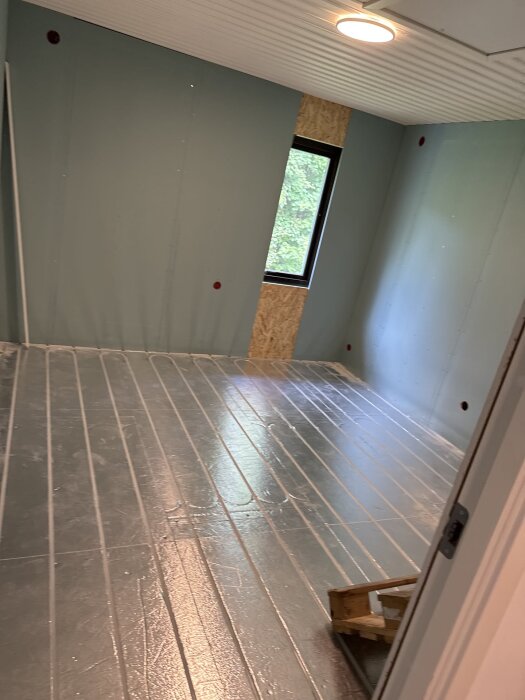 Rum under renovering med golvvärmeslangar på golvet, gipsade väggar och ett fönster.