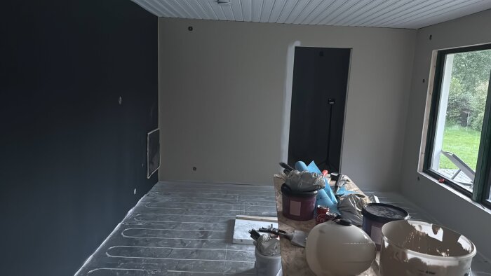 Rum under renovering med isolering, målad vägg i svart och vitt samt utrustning och material på golvet, inklusive en Coca-Cola-burk.