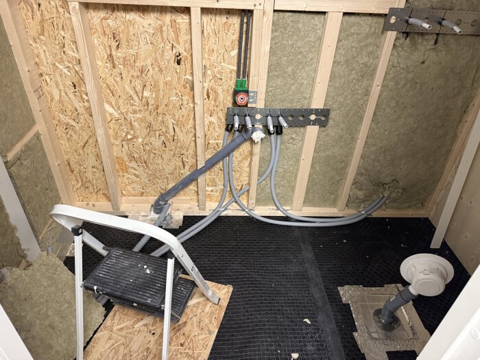 Badrum under renovering med isolerade väggar, VVS-rördragning och byggmaterial förberedda för golvvärme och plywoodväggar. Stege står framför.