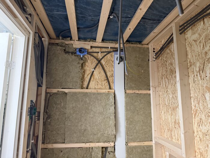 Isolerad vägg under renovering med synliga träreglar och installationsrör i ett utrymme, troligen ett badrum under konstruktion.