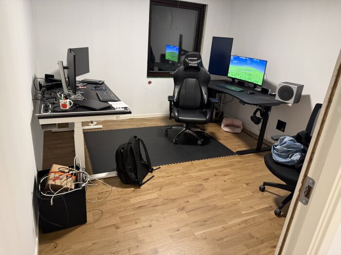 Ett rum med två skrivbord, datorer och en kontorsstol. Högtalare och kablar syns, samt en mugg med pennor. Trägolv med en svart matta.