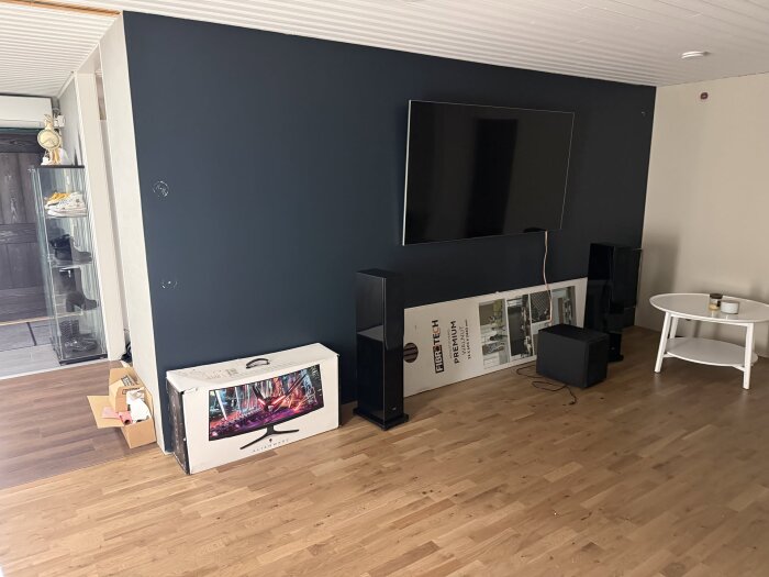 Rum med nyrenoverad mörkblå vägg, väggmonterad TV, högtalare, ett runt bord, och en oöppnad tv-låda på parkettgolv.