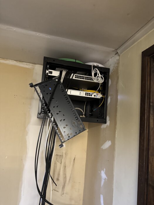 Vägghängt serverrack med nätverkskablar och utrustning, delvis öppet, mot en delvis målad innervägg.