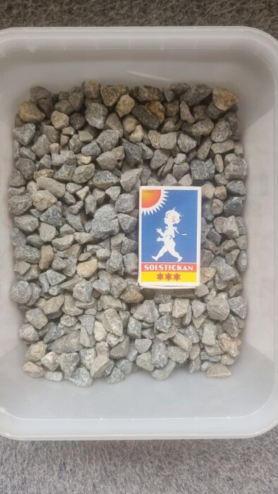 Små stenar i en plastbehållare bredvid en tändsticksask med etiketten "SOLSTICKAN".