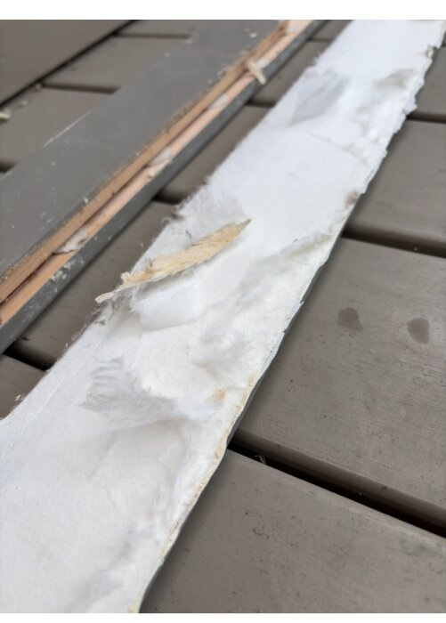 Pappersliknande material med lurvig yta bakom golvsockel, misstänkt asbest eller gammal tapet, ligger på grått plankgolv.