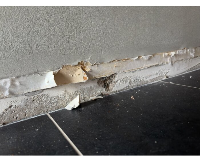 Lurvigt, pappersliknande material bakom golvsockel mot en vägg i en hall. Materialet ifrågasätts för eventuell asbestinnehåll.