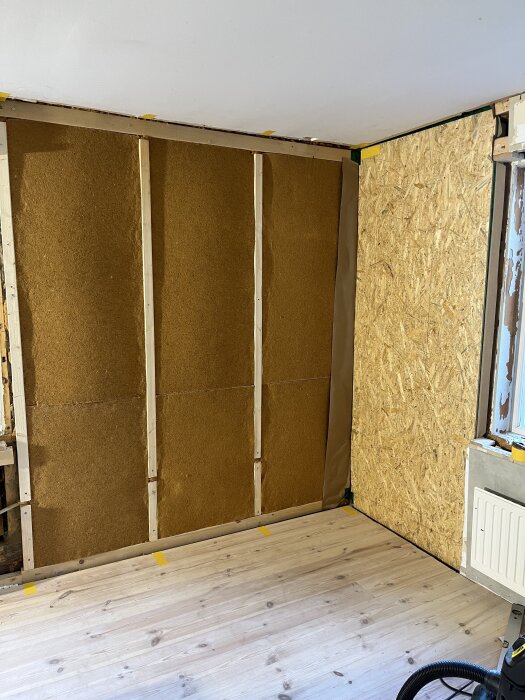 Vägg under arbete med isolering, ångbroms och OSB-skiva. Golv av trä, rumshörn visar skarv med överlappande ångbroms och tejp.