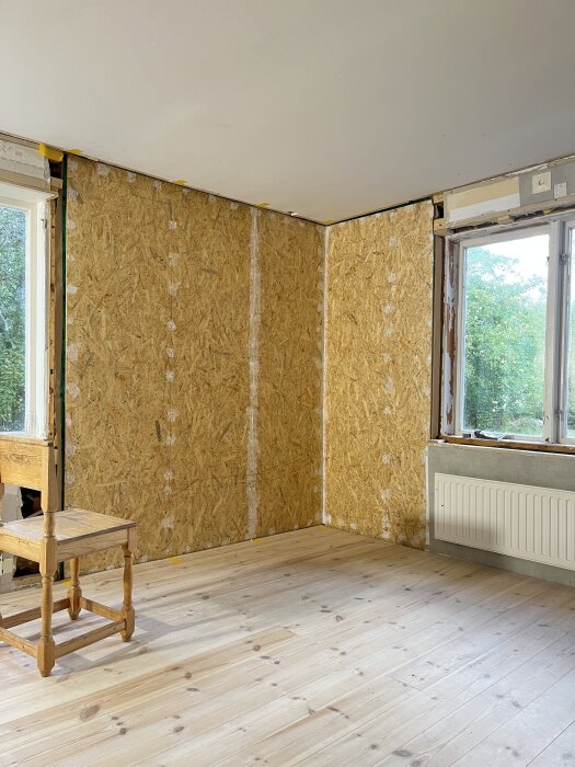 Rum under renovering med osb-skivor på väggarna, obehandlat trägolv och en stol. Två fönster ger utsikt mot grönska utanför.