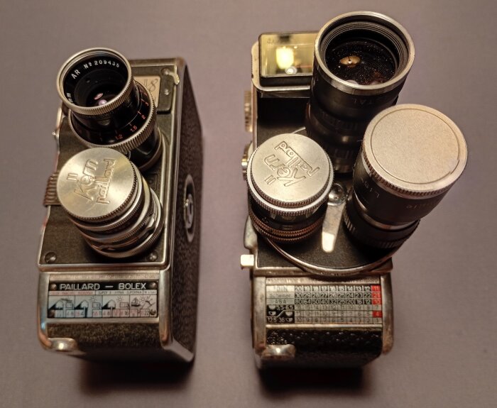 Två vintagedubbelåttakameror från Paillard Bolex, en med revolverobjektiv, båda med fjäderverk, sedda uppifrån mot en grå bakgrund.
