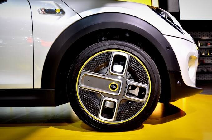 Närbild på en bilfälg med en unik design och luftreflektorer, monterad på en Mini Cooper parkerad på ett gult golv.