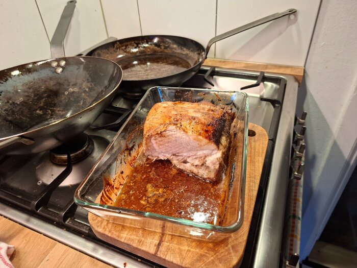 Stekt köttbit i en glasform på en spis, omgiven av två stekpannor.