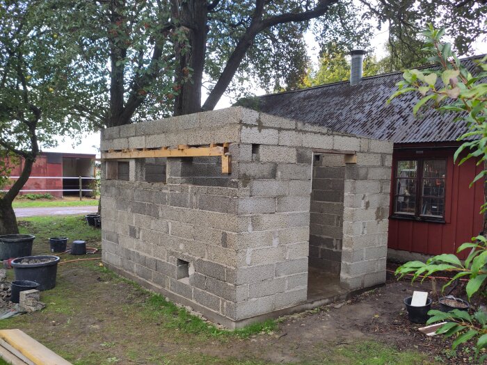 Byggnad under konstruktion med väggar av betongblock bredvid ett rött träskjul, omgiven av träd och grönska.