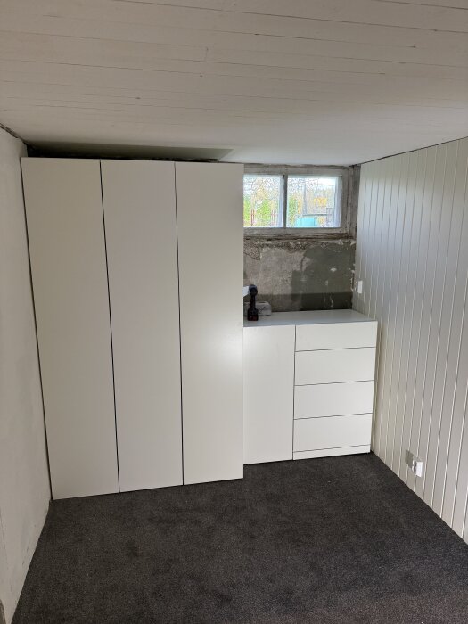 Vit garderob och byrå i ett källarrum med vitmålad takpanel och grå matta. Fönster med utsikt mot trädgård.