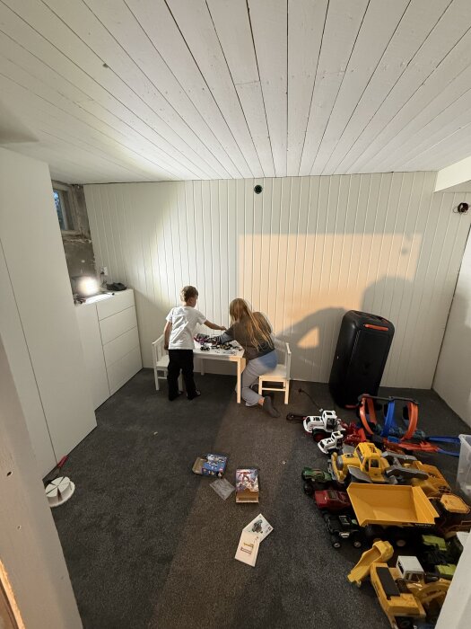 Barn leker med leksaker i ett källarrum med vit träpanel och grå matta. Rummet har leksaker och ett mindre bord med stolar.