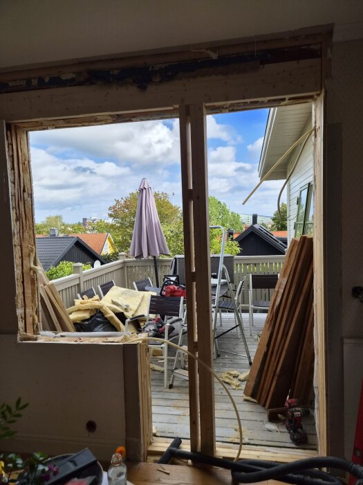 Öppning för nytt fönster och altandörr i byggnadsväggen, träplankor och isolering synliga, veranda utanför med stolar och bord.