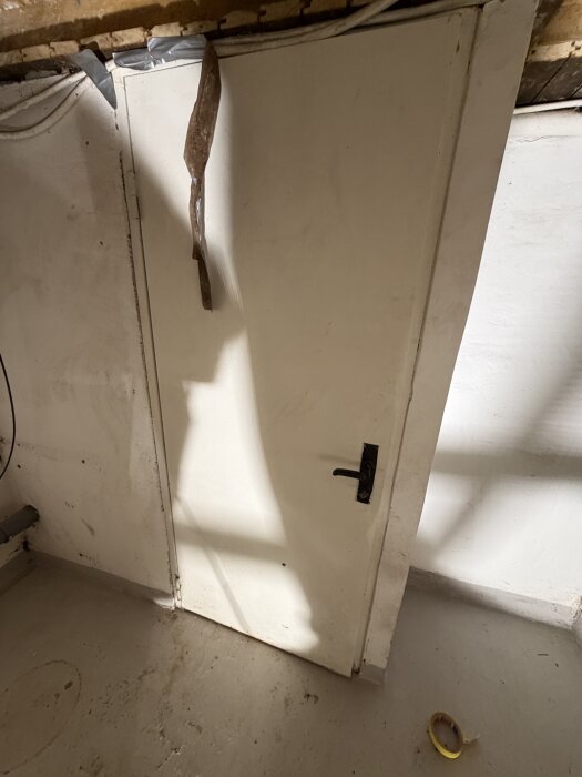 En vit innedörr med svart handtag, skuggad av ljus, ligger an mot en ljus vägg. Dörren är trasig upptill och tejpad.