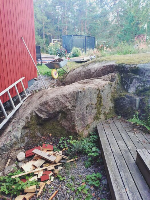 Bergknalle vid röd byggnad med en hög av träbitar och spade intill. Trädäck till höger och skottkärra i bakgrunden nära trädgård.