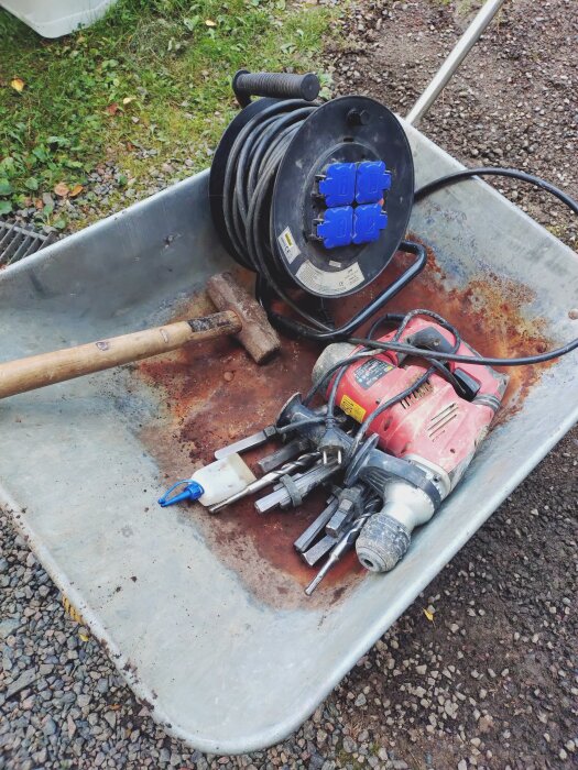 Verktyg i en skottkärra: borrmaskin, slägga, borr och elkabel på grus, med grön mark i bakgrunden. Används för att bearbeta berg.