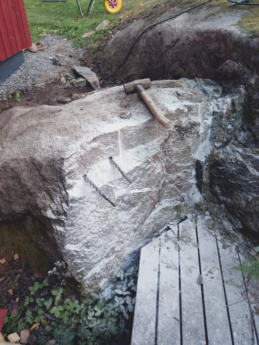 Hammare ligger på en delvis bearbetad bergknalle nära en liten träplatta, surret av sprickor syns på stenen.